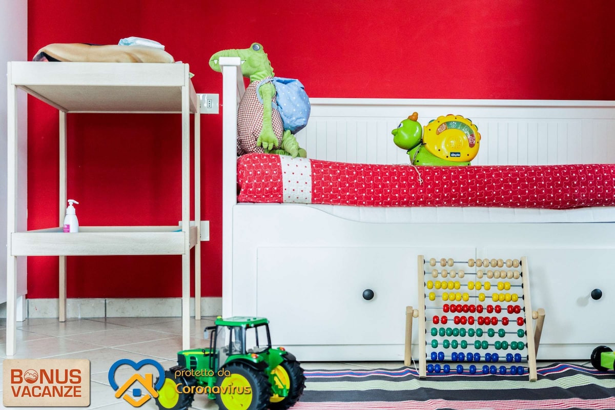 A children's bedroom is visible, featuring a red wall backdrop. A white bed with a red patterned cover is seen, adorned with soft toys and a colorful clock. A wooden play table is present alongside a counting board, while a toy tractor completes the playful atmosphere.