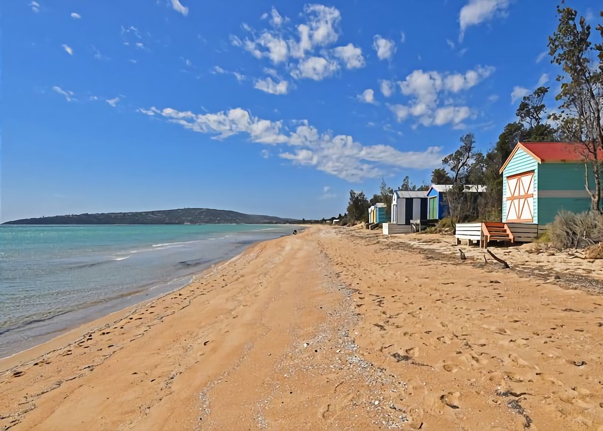 A stretch of sandy beach is seen, bordered by colorful beach huts in varying styles. Gentle waves lap at the shore under a clear blue sky, while trees line the background, providing a serene and tranquil coastal environment.