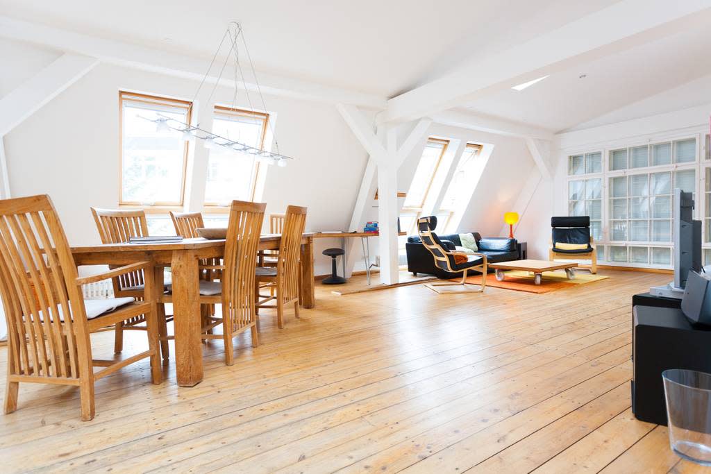 The spacious living area features a wooden dining table with six chairs, alongside a comfortable seating arrangement that includes a leather sofa. Bright natural light filters through multiple windows, highlighting the wooden floor and the unique architectural details of the loft.
