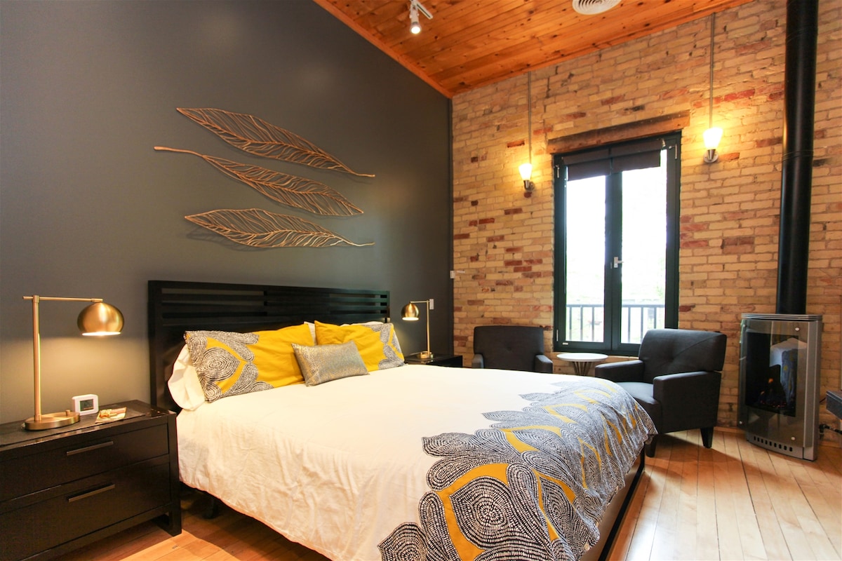 A contemporary room features a king size bed adorned with patterned linens and accented by a decorative wall art of leaves. Two armchairs are positioned next to a window, while a gas fireplace adds warmth. The ceiling showcases warm wooden beams.