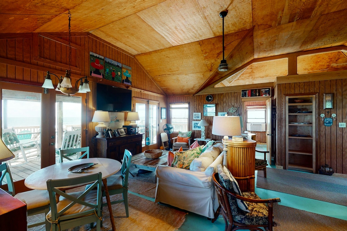 The living area features high, wooden ceilings that enhance the spacious feel. Various seating options are arranged around a central coffee table, with light streaming through multiple windows. A dining table is positioned nearby, complementing the rustic decor and beachy ambiance.