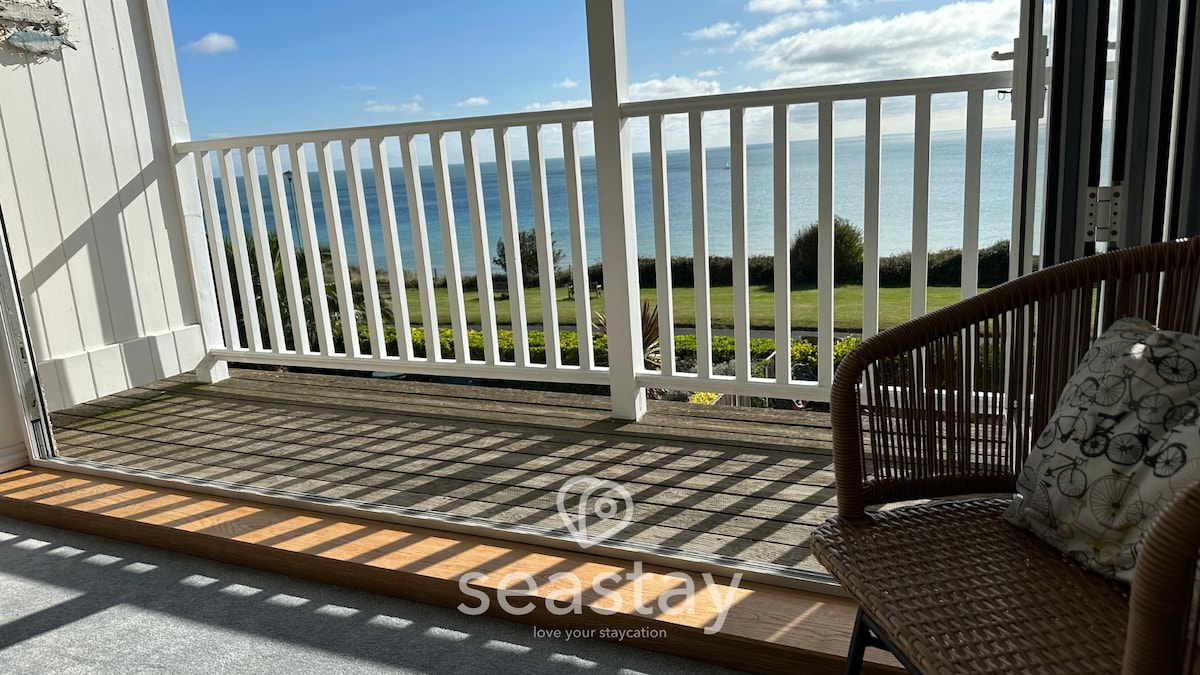 A private balcony showcases a serene view of the English Channel, with the sun casting patterns on the deck. A comfortable wicker chair is positioned to enjoy the tranquil atmosphere, while the calming hues of the sea extend in the background.