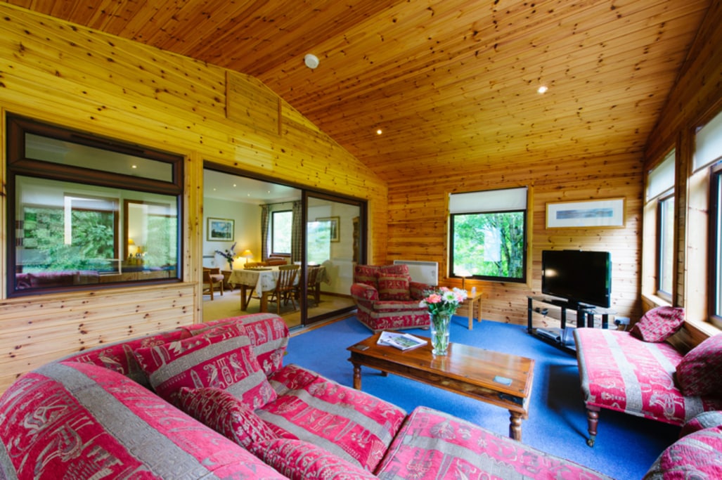 The living area features wooden paneled walls and an open layout, complemented by large windows allowing natural light. Comfortable seating arrangements include a sofa and chairs adorned with patterned upholstery. A coffee table sits in the center, while a television is positioned nearby.