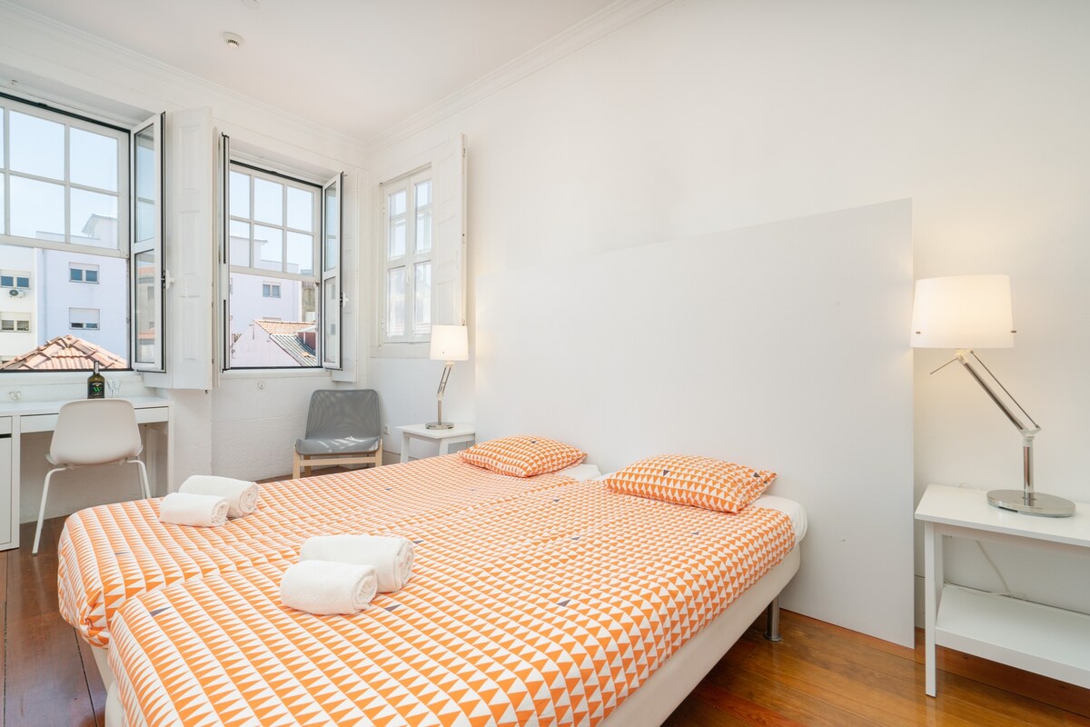 The bright bedroom features three single beds dressed in vibrant, patterned linens. Natural light filters through two open windows, illuminating the minimalist decor. A chair and a small table are positioned nearby, with bedside lamps providing additional lighting.