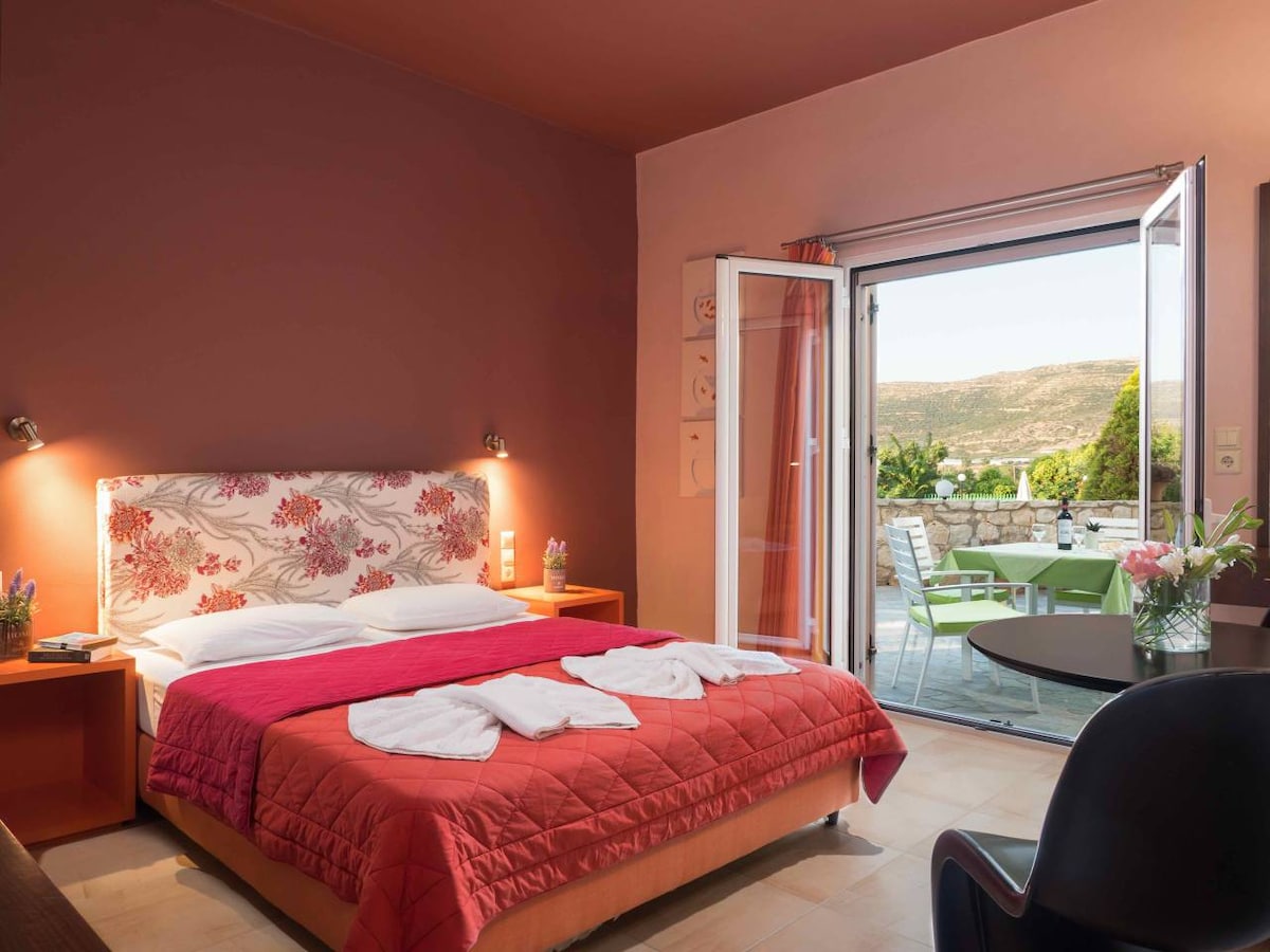 A cozy room with a red bedspread is complemented by a floral-patterned headboard. Large glass doors open to a private balcony with a view of the garden. A round dining table and dark chair are present, enhancing the inviting atmosphere.