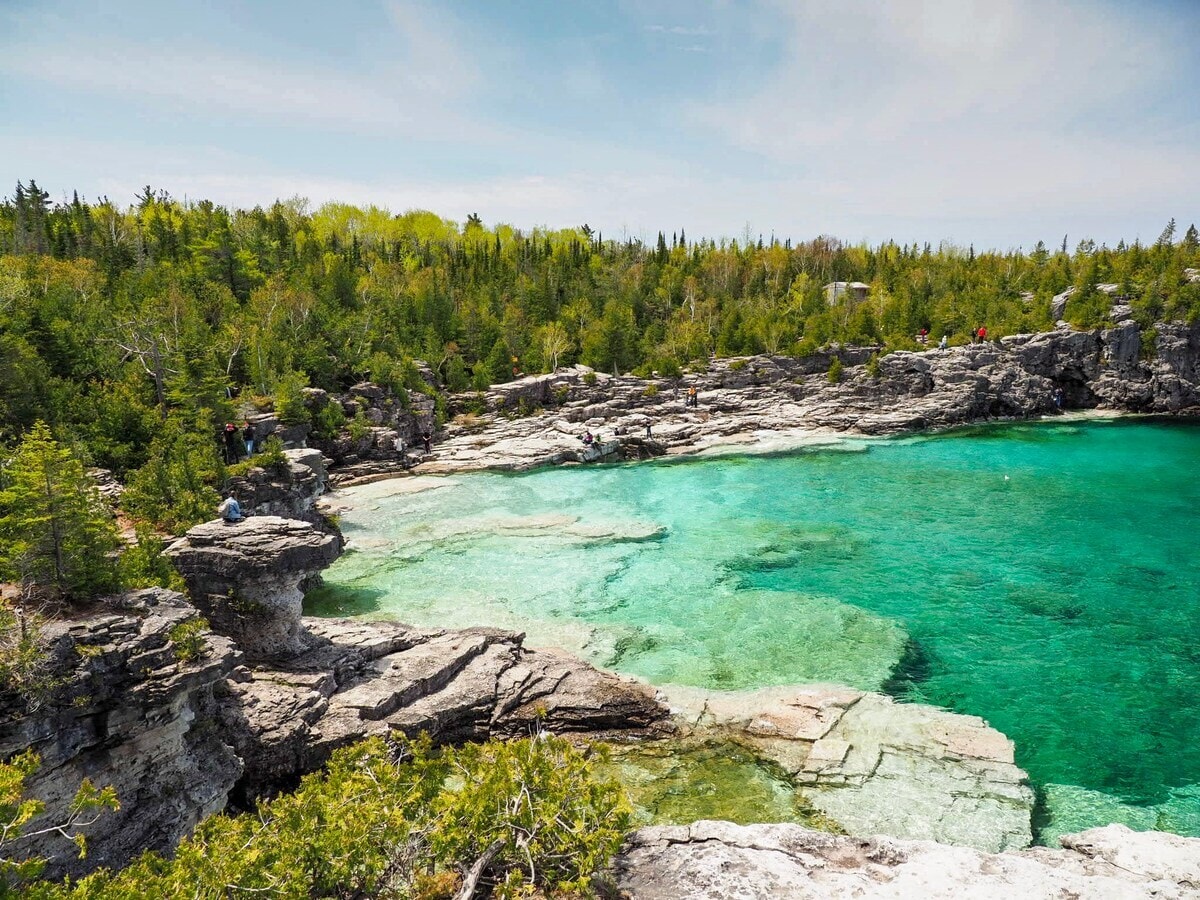 A rocky shoreline sits along the edge of a clear turquoise body of water, framed by lush greenery. The scene captures a blend of rugged stone formations and vibrant trees, suggesting opportunities for exploration and relaxation in nature.