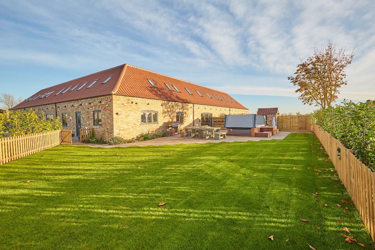 A spacious rural barn is viewed from the beautifully maintained garden, featuring a large grassy area bordered by a wooden fence. The exterior combines natural stone with a red tiled roof, while the patio area includes inviting seating and a hot tub.