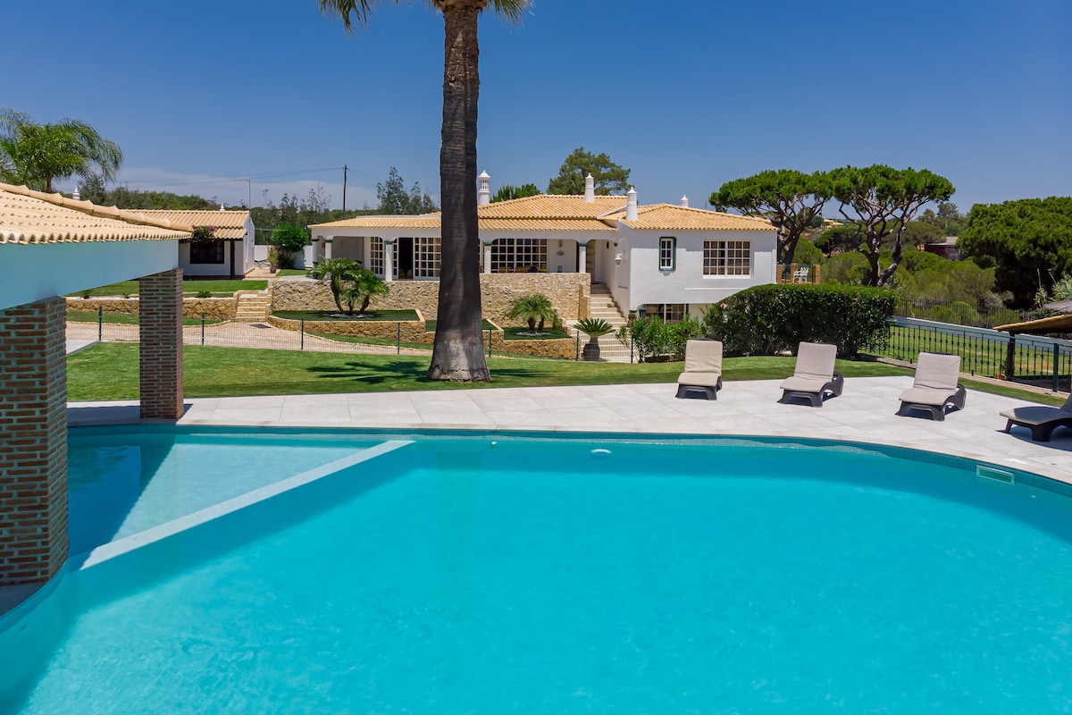 A large swimming pool is featured in the foreground, with clear blue water reflecting sunlight. Several lounge chairs are arranged around the pool area. In the background, a spacious villa can be seen, surrounded by lush green grass and landscaped gardens.