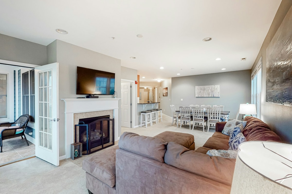 An open-concept living area features a spacious sectional sofa in a soft shade, complemented by a coffee table. A gas fireplace is located beneath a mounted TV, while a dining area with a large table and chairs is visible in the background. Natural light fills the room from nearby windows.