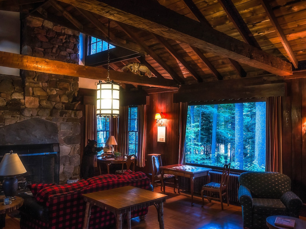 The living area features a high, beamed ceiling with wooden accents, stone fireplace, and large windows framing views of the surrounding trees. Comfortable seating options include a plaid sofa and an armchair, while a wooden table and chairs provide a dining space.