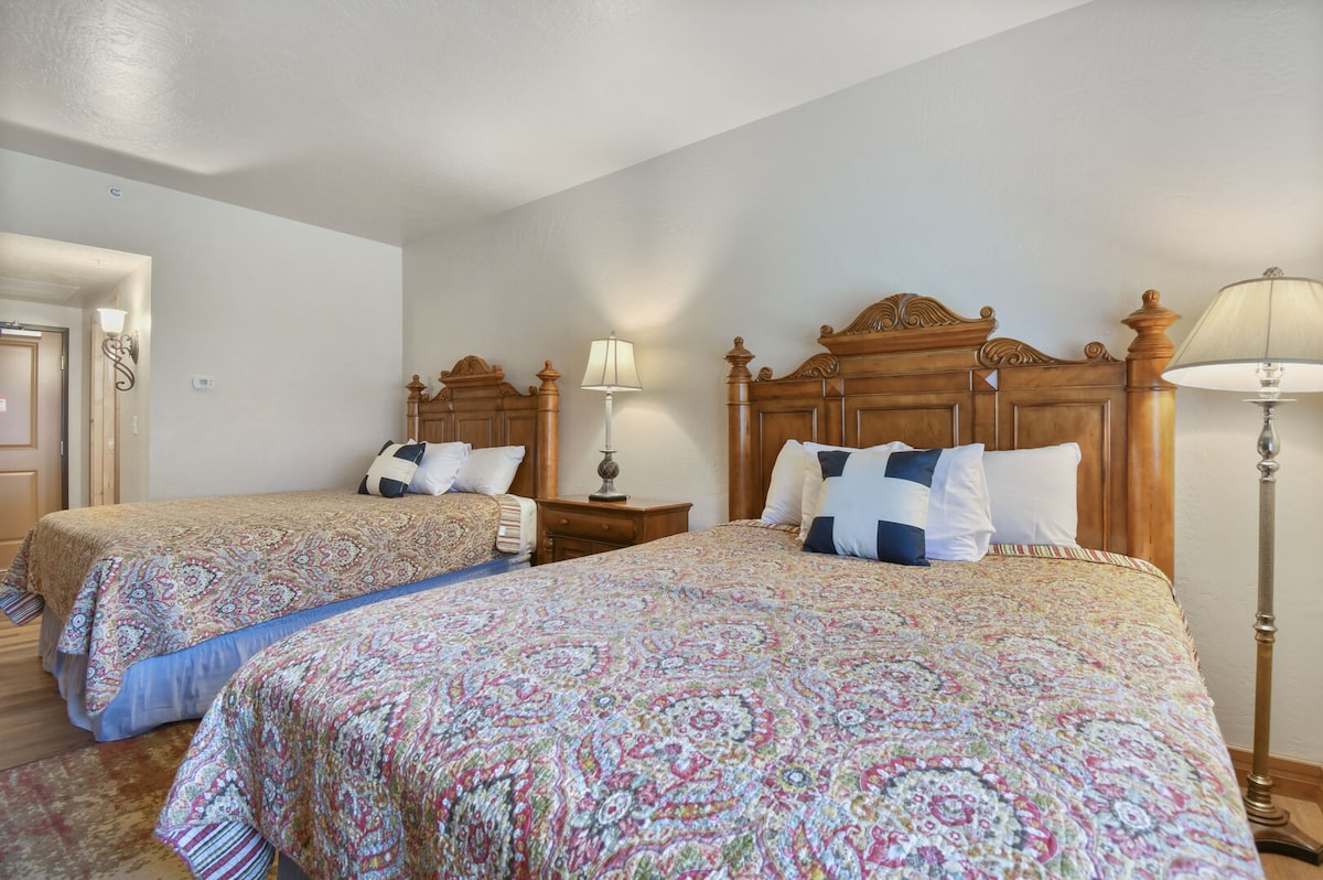 A comfortable sleeping area is presented with two wooden queen beds, each adorned with decorative quilts in warm colors. A wooden nightstand, fitted with a lamp, is positioned between the beds. Soft light is emitted by the lamp, enhancing the inviting atmosphere.