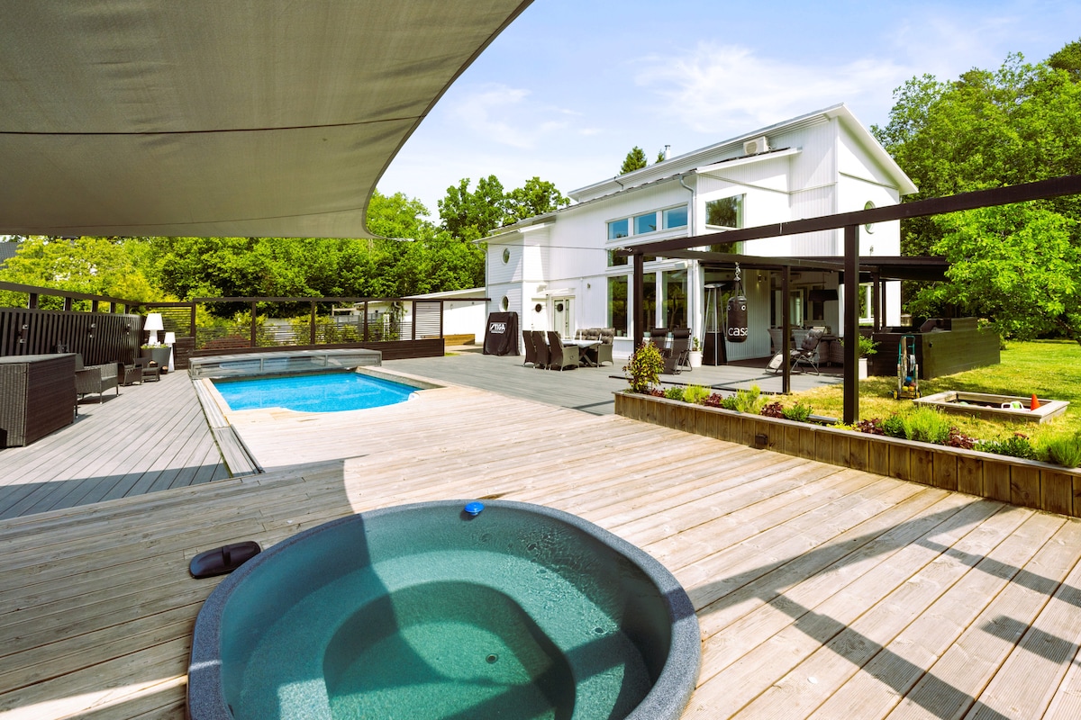 An outdoor area showcases a heated pool and a hot tub surrounded by a wooden deck. Comfortable lounge furniture and a dining set are visible, creating a space for relaxation. Lush greenery borders the property, enhancing the tranquil atmosphere.
