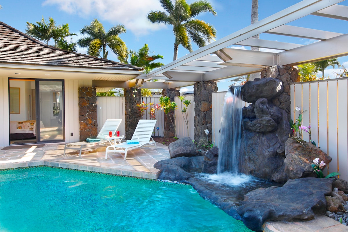 A private pool area is showcased, featuring a serene waterfall cascading over natural stones. Two lounge chairs are positioned nearby, inviting relaxation amidst lush tropical landscaping. An open trellis above allows for ample sunlight, while the exterior of the home is visible in the background.