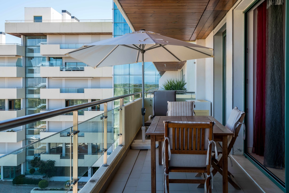 An outdoor balcony area features a wooden dining table with matching chairs and a large umbrella providing shade. A partial view of the surrounding contemporary buildings is visible in the background, with greenery enhancing the tranquil setting.