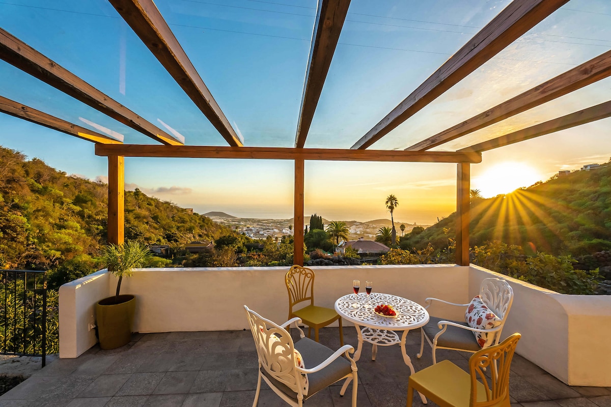 A covered terrace features a round table with decorative white chairs. The setting is enhanced by a scenic view of the hills and sea, illuminated by the warm glow of the setting sun. Lush greenery is visible in the background.
