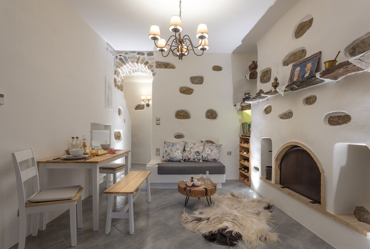 A cozy sitting area is depicted with a contemporary dining table and bench, complemented by a small round coffee table. Natural stone accents embellish the walls, while a traditional fireplace adds warmth. Soft textiles are arranged on the floor, contributing to a welcoming ambiance.