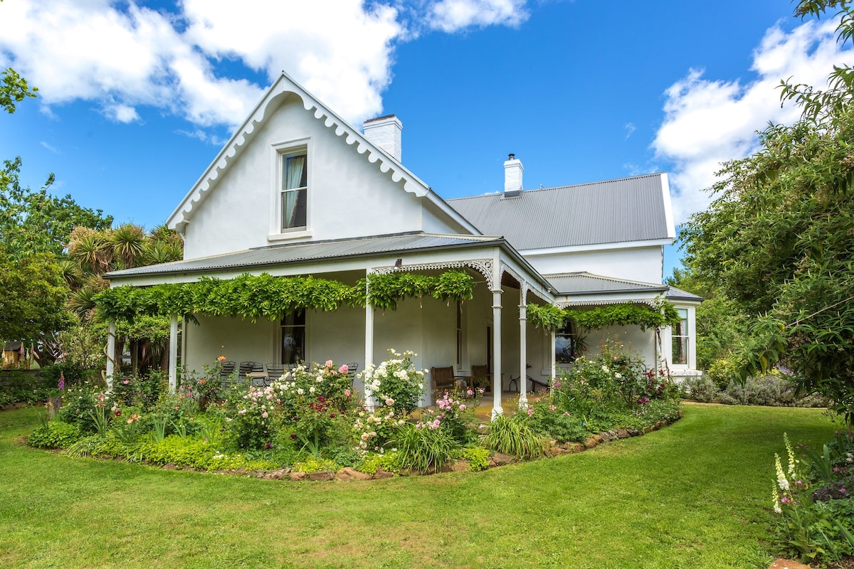 A charming two-story farmhouse is surrounded by lush gardens and greenery. The porch features vine-covered posts, offering seating areas for relaxation. Bright flowers bloom throughout the landscaped yard, creating a serene outdoor setting under a clear blue sky.