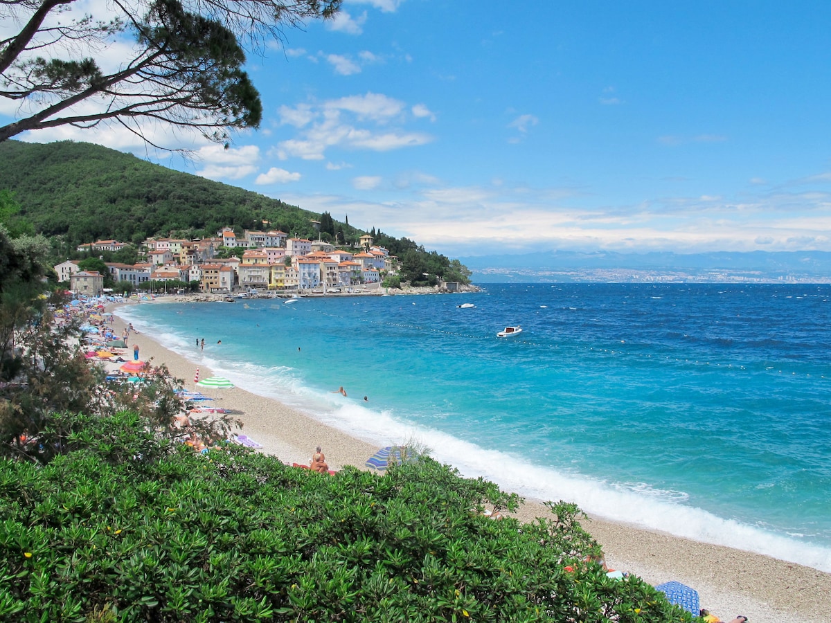A sandy beach is depicted with gentle waves lapping at the shore. Colorful sun loungers and umbrellas can be seen along the coastline, while a hillside village with various buildings overlooks the sea. The vibrant blue water contrasts with the greenery in the foreground.