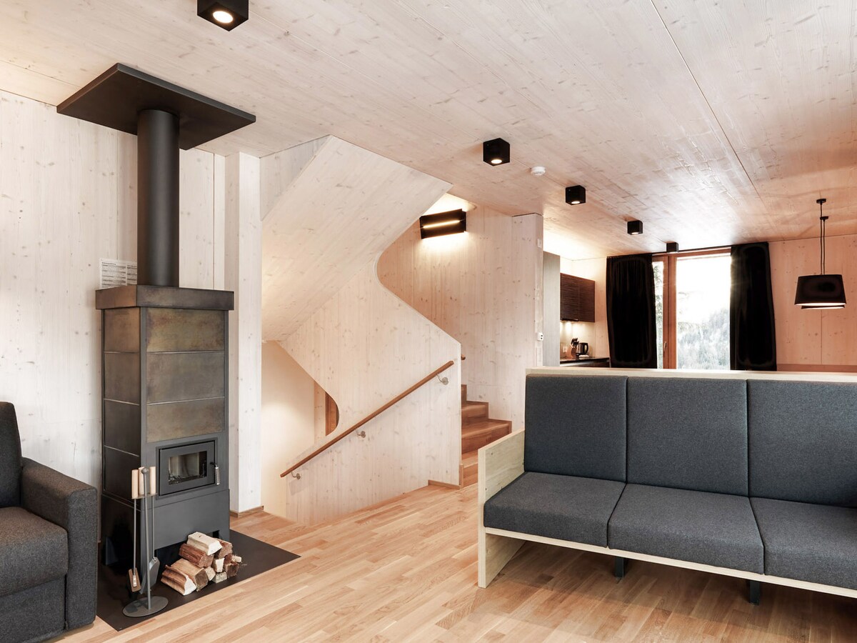 The modern living room features a wood-burning stove adjacent to a comfortable gray sofa and a minimalist seating area. Natural wood paneling creates a warm ambiance, enhanced by soft lighting fixtures. A staircase leads to the upper level, while large windows provide views of the outdoors.
