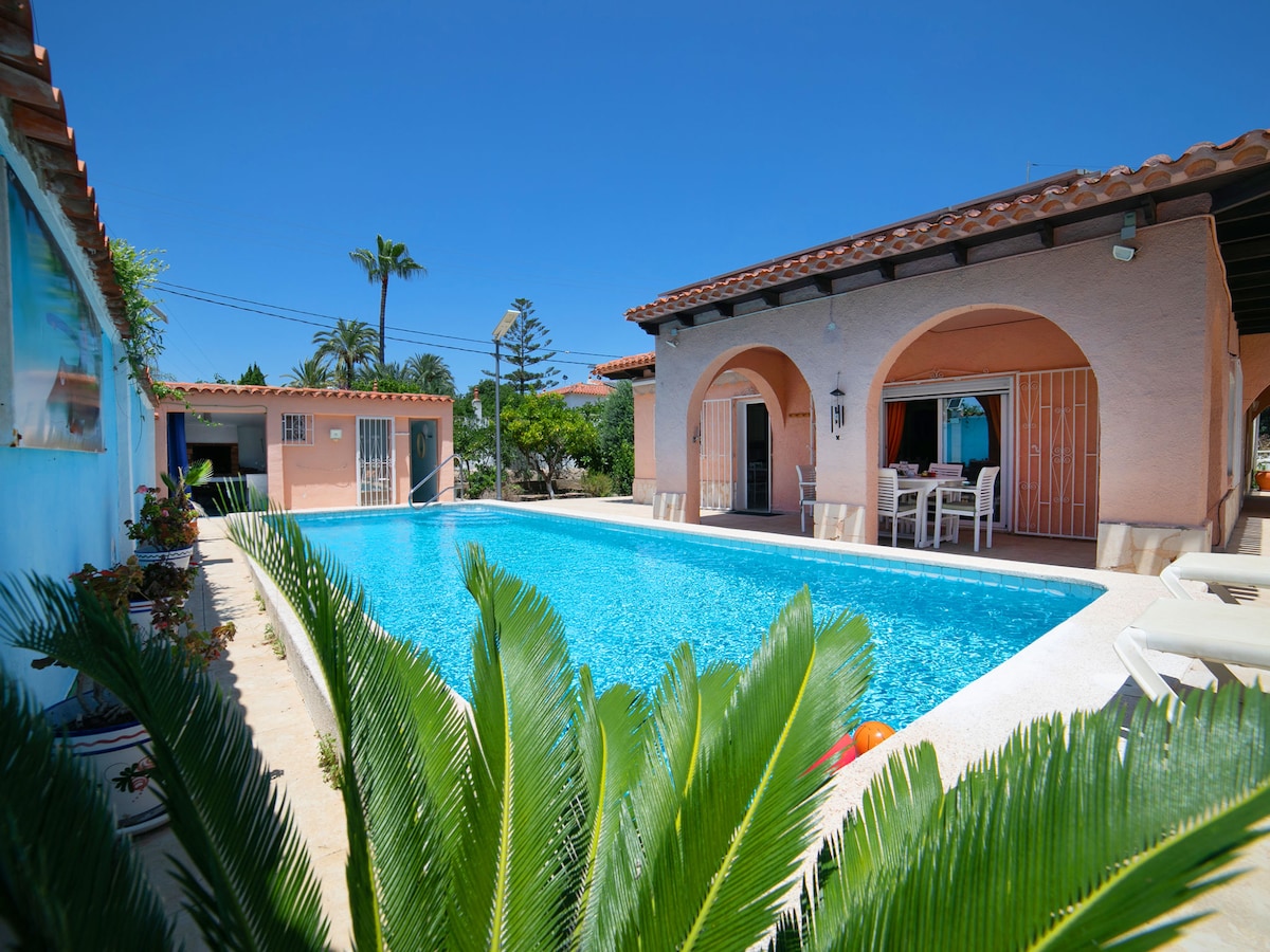 A large swimming pool is framed by a smooth deck with sun loungers and a dining area, surrounded by lush greenery. The house features a warm exterior and arched doorways, blending harmoniously with the inviting outdoor space.