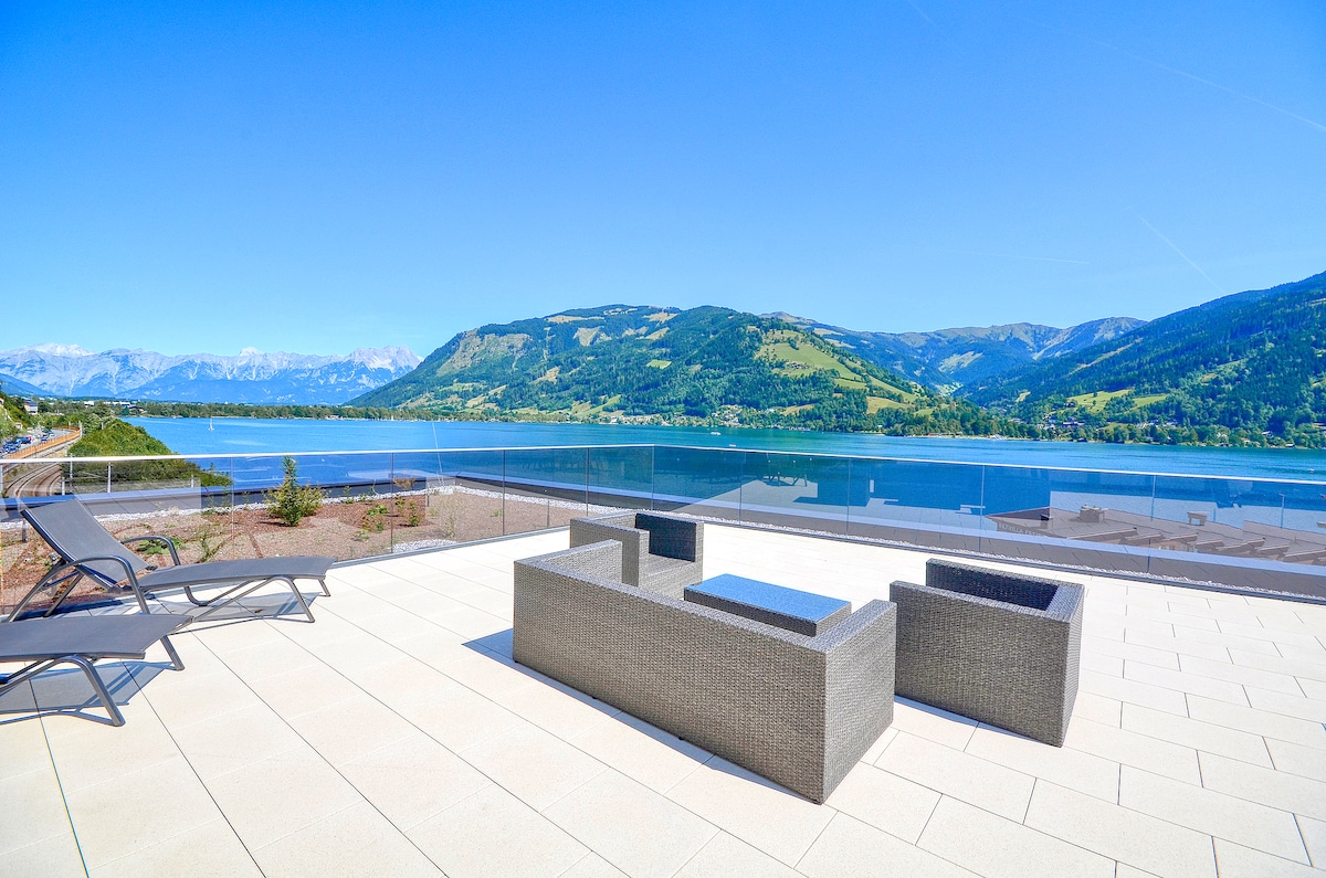 A spacious roof-top terrace features a modern seating arrangement with a coffee table, offering views of Lake Zeller and surrounding mountains. Lounge chairs are positioned for relaxation, while the sun illuminates the outdoor space, enhancing the tranquil atmosphere.