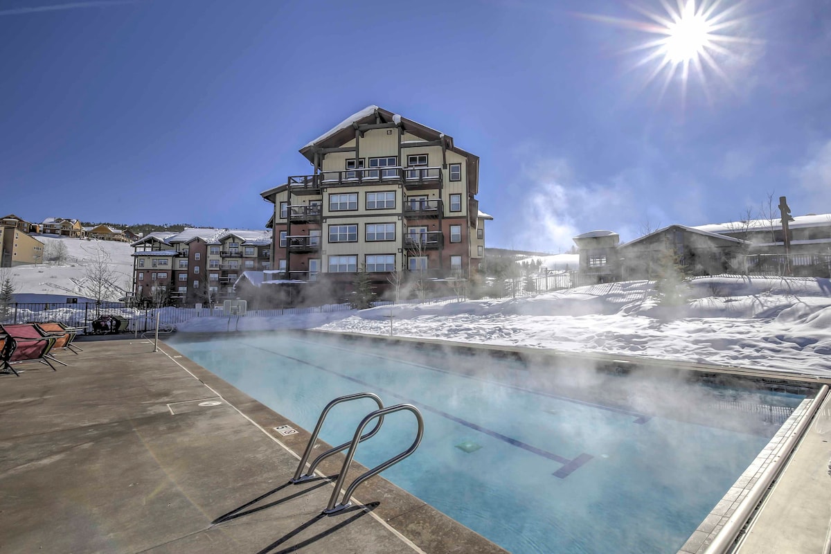 An outdoor heated pool is visible surrounded by snow-covered landscapes. The pool features metal ladders leading into the water, with steam rising gently in the chilly air. A multi-story building stands in the background under a clear blue sky.