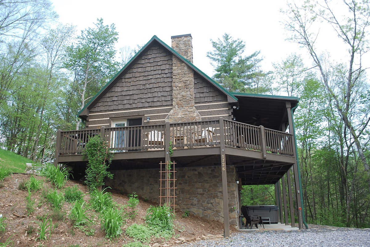 A log cabin is nestled among trees, showcasing a wrap-around deck with ample seating. The stone chimney adds rustic charm, while the covered area extends the living space outdoors. A hot tub is visible on the lower patio, surrounded by landscaped greenery.