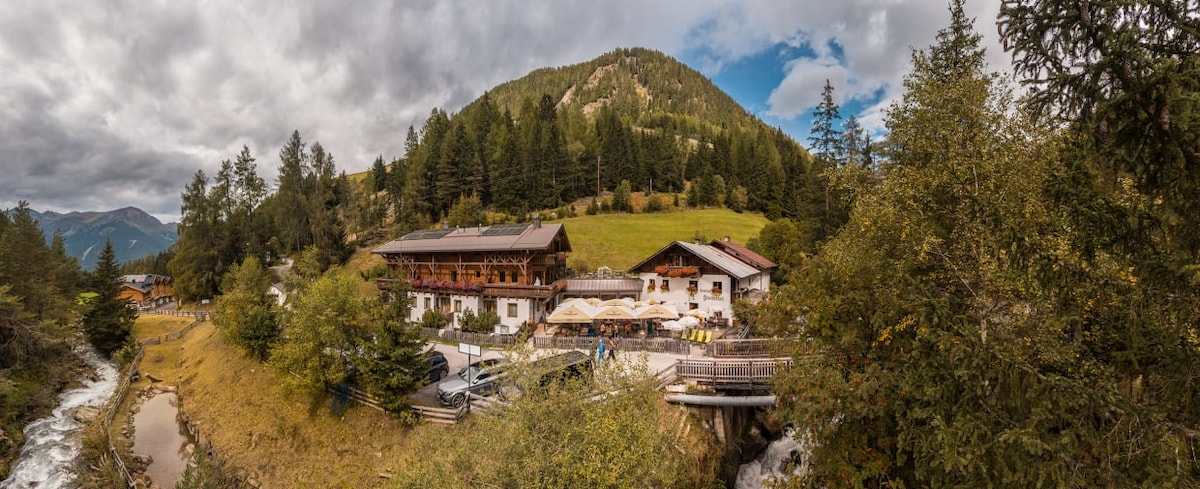 A panoramic view of a mountain hotel is showcased, nestled among lush greenery and a gentle slope. A flowing river is visible in the foreground, while the surrounding mountains rise in the background under a partly cloudy sky.