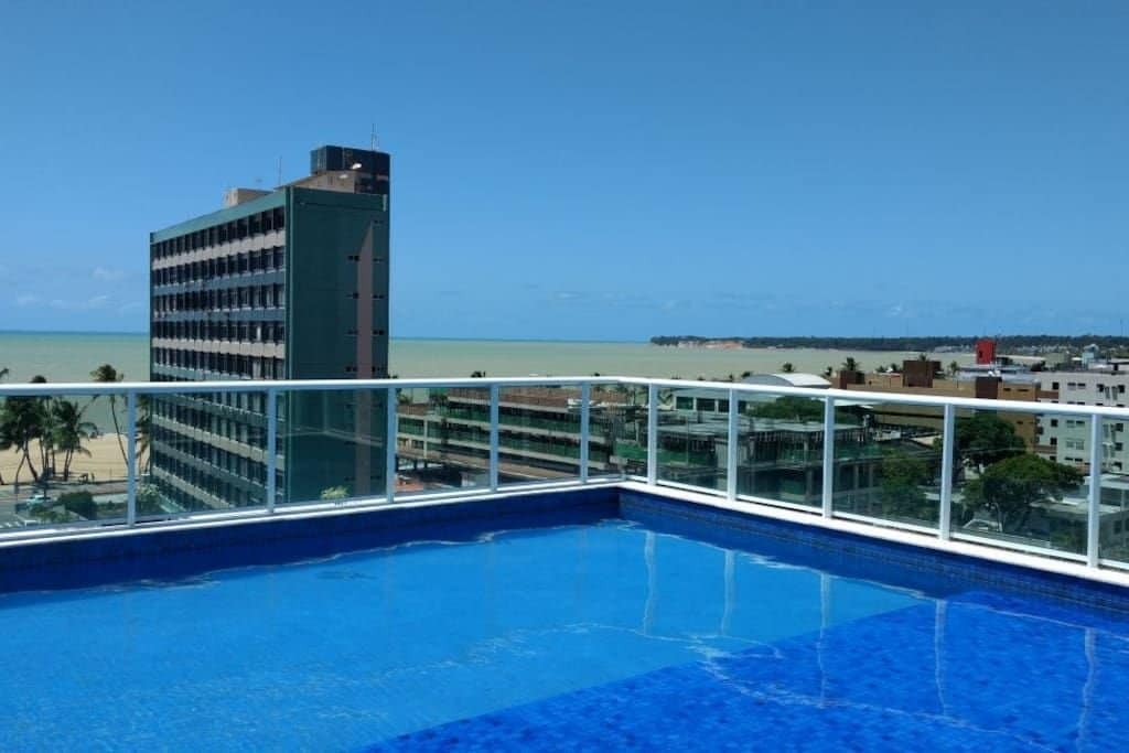A rooftop pool is featured, surrounded by a glass railing that offers unobstructed views of the coastline and the cityscape. The water is a vibrant blue against the clear sky, creating a serene setting for relaxation.