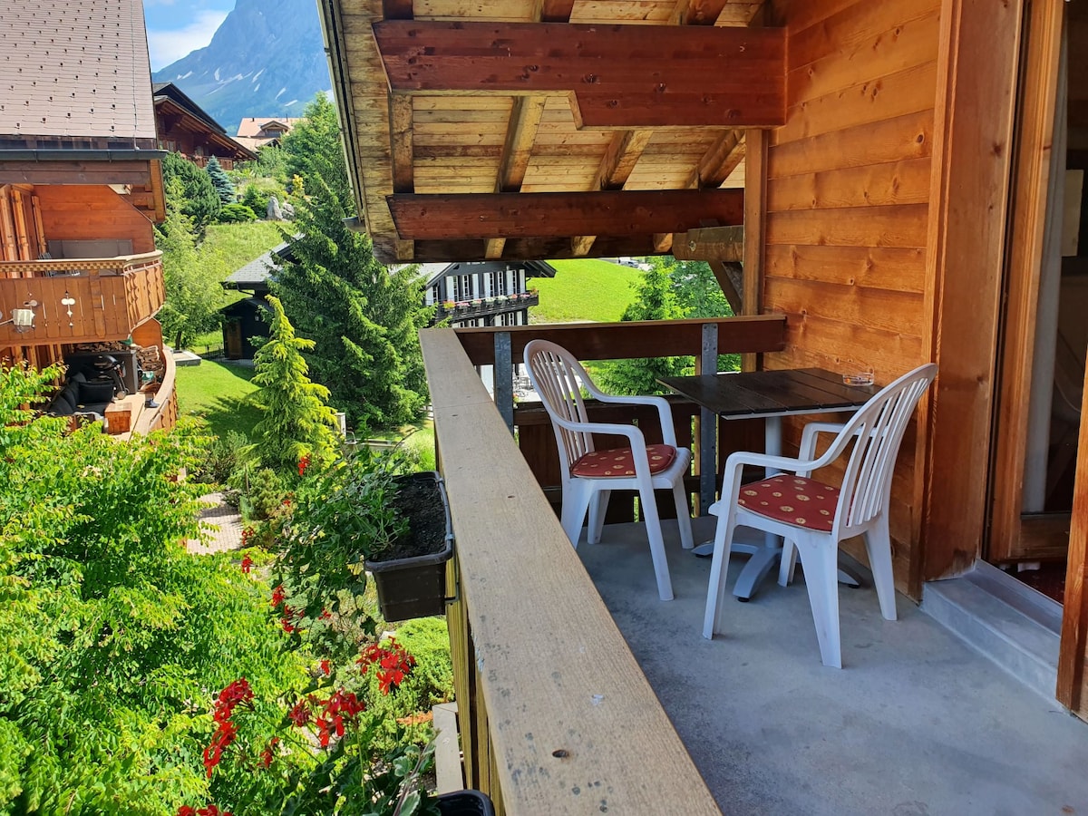 A balcony features a wooden railing with two white chairs and a small table. Nearby, vibrant greenery and flowers can be seen, providing a natural backdrop. The slopes of the mountains are visible in the distance under clear skies.