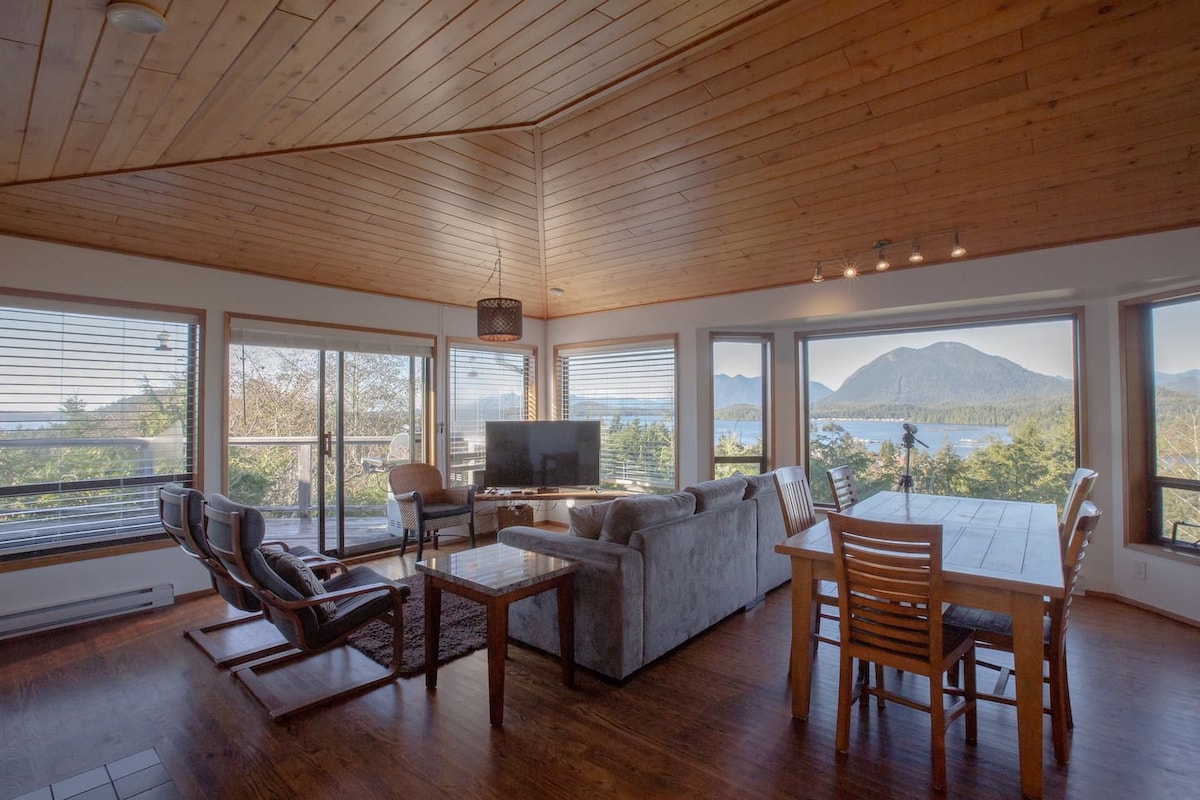 An open concept living and dining area is showcased, featuring large windows offering expansive views of the surrounding landscape. A comfortable gray sofa faces a flat-screen television, while a wooden dining table accommodates multiple guests. Natural light fills the space, highlighting the wooden ceiling beams.