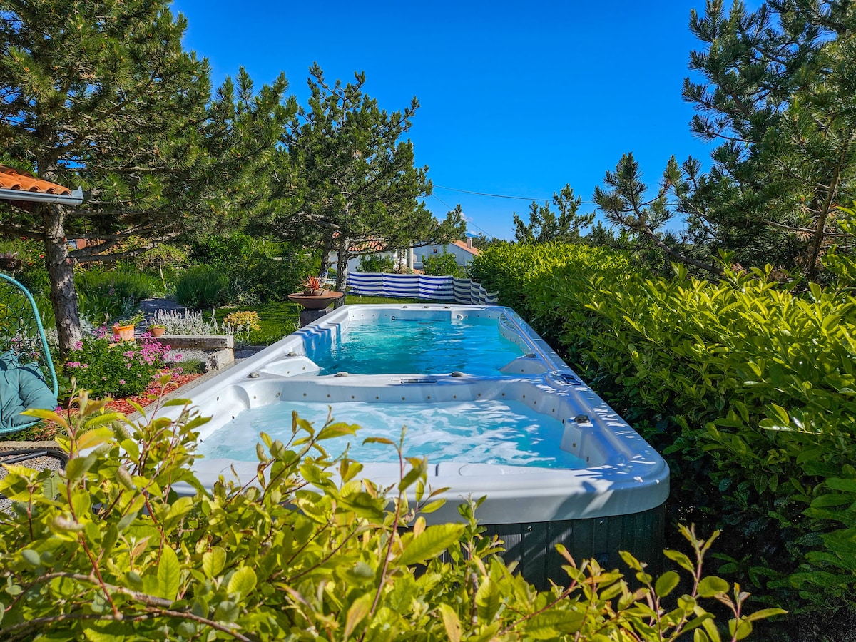 A seasonal swimming pool is surrounded by lush greenery and colorful flowers. The clear blue water is inviting, while a patio area with loungers is visible in the background. Tall pine trees provide shade and enhance the serene outdoor setting.