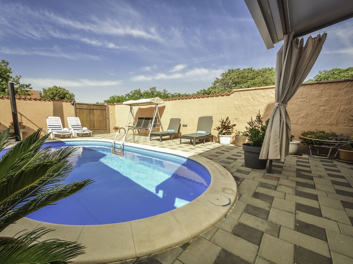 A private swimming pool is surrounded by a sun terrace, featuring two loungers and a swing. Potted plants are positioned around the area, and a shaded seating space is available. The pool's clear water reflects the blue sky, creating a relaxing outdoor environment.