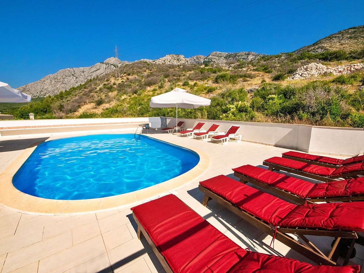 A circular swimming pool is surrounded by several sun loungers with red cushions. Bright blue water is visible in the pool, and an outdoor area features light-colored tiles. Umbrellas provide shade nearby with a hilly landscape in the background under clear blue skies.