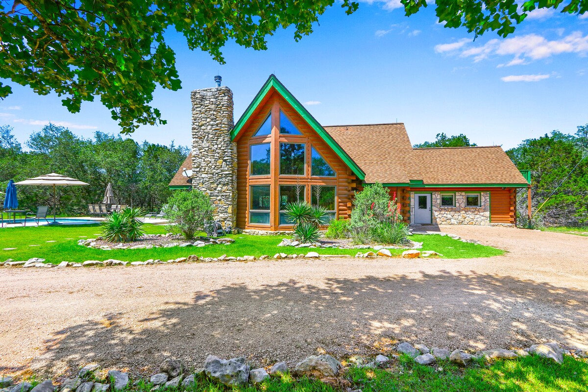 A Texas-style log cabin is presented with a stone chimney and a prominent angled roof. Floor-to-ceiling windows allow natural light to illuminate the interior. Surrounding landscaping features pathways, shrubs, and an outdoor seating area near the pool, enhancing the rustic charm.