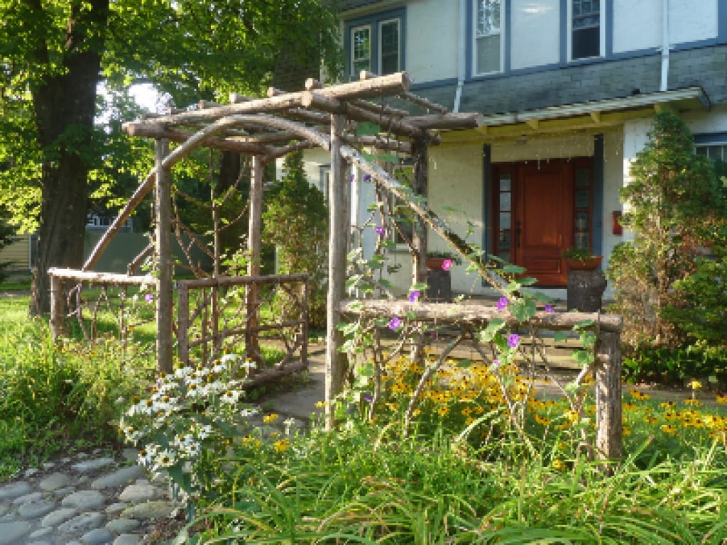 A rustic wooden arbor adorned with climbing vines and colorful flowers frames the entrance. Surrounding the path, lush greenery and seasonal blooms create a welcoming atmosphere. The classic colonial house is visible in the background, with its wooden door highlighted by natural light.
