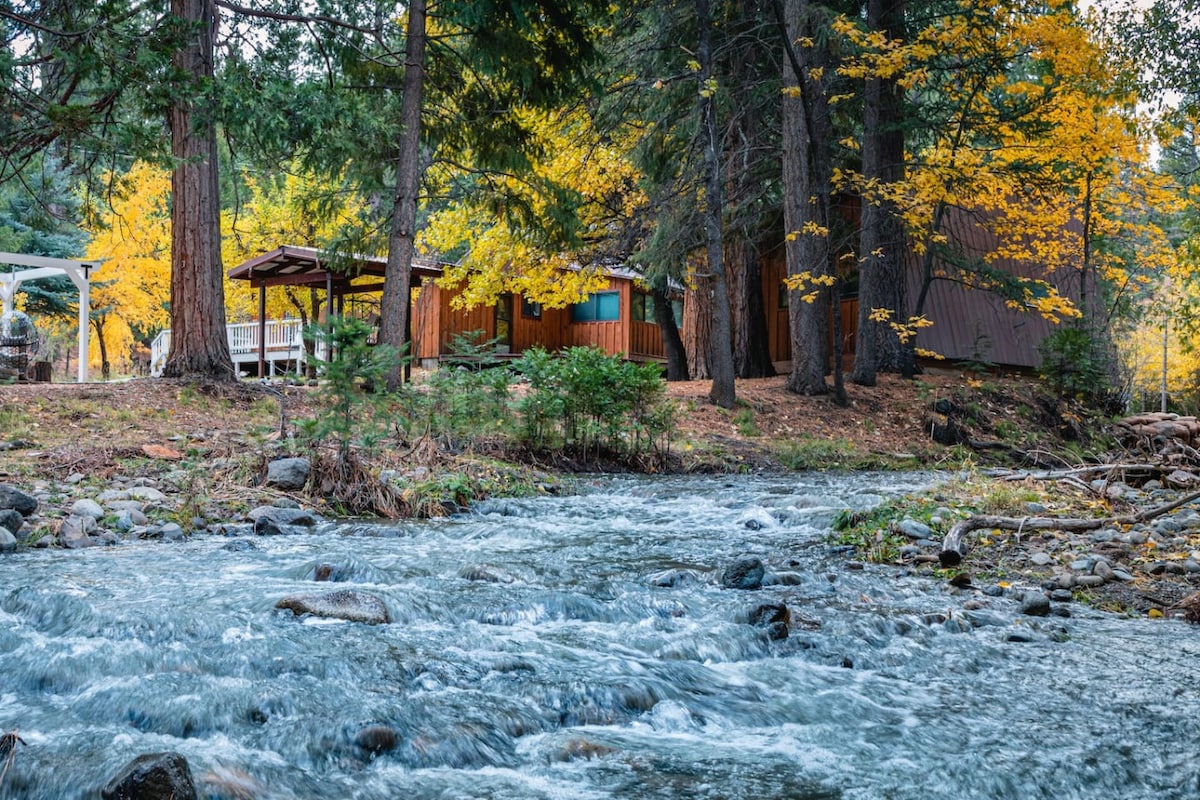 A serene creek flows through the foreground, bordered by smooth stones. A wooden cabin is nestled among tall trees, showcasing autumn foliage. An outdoor seating area can be seen nearby, offering a peaceful spot to enjoy the surroundings.