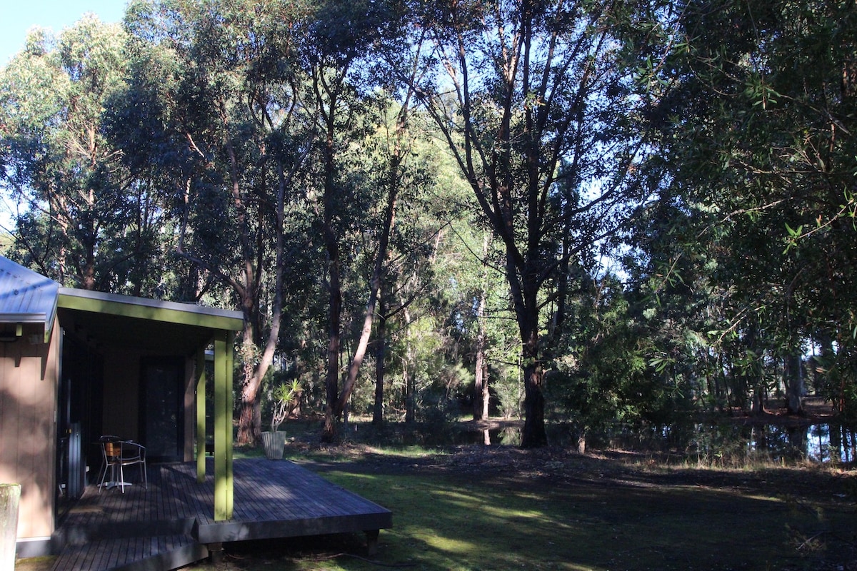 A spacious cottage is situated near a wooden deck, surrounded by tall trees. Sunlight filters through the foliage, creating a serene atmosphere. The natural landscape includes a reflective body of water, enhancing the tranquil setting.