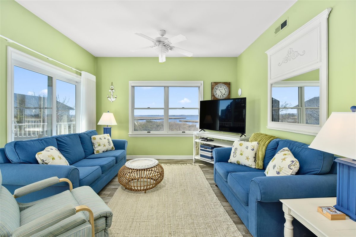 A bright living area showcases two blue sofas adorned with decorative pillows, accompanied by a light-colored armchair. A wooden coffee table is centered on a light rug, while large windows provide a scenic view of the bay. A television is positioned on a white cabinet.