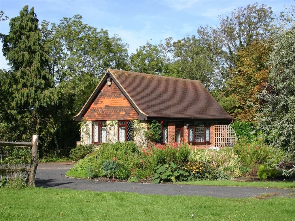 A charming cottage features a combination of wooden and stone exterior, surrounded by lush greenery and colorful flowerbeds. Spacious garden areas provide an inviting atmosphere. Large windows with ornate patterns allow natural light to fill the interior, enhancing the cozy appearance of the building.