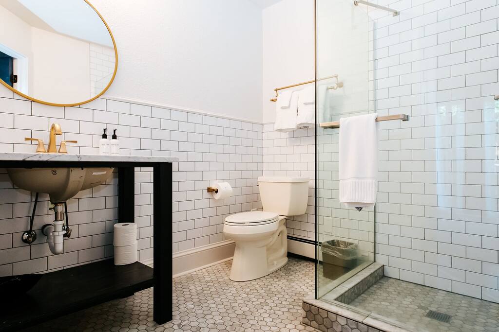A clean and modern bathroom is highlighted by white tiled walls and a glass shower enclosure. A round mirror reflects soft light above a dark, minimalist vanity with a sink. Essential toiletries are neatly arranged, and a toilet is positioned beside the shower.