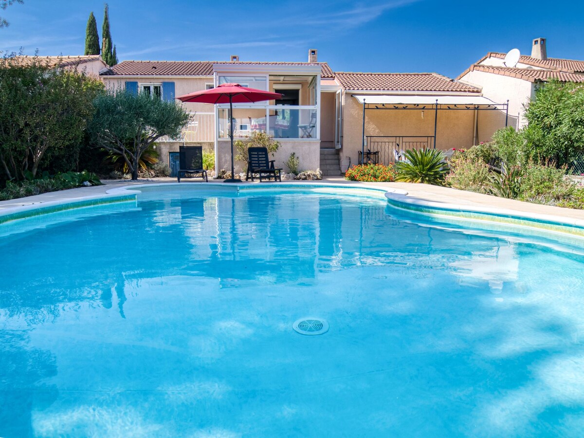 A clear blue swimming pool is surrounded by well-maintained gardens. A deck with sun loungers is visible, accompanied by a large umbrella for shade. The house is showcased in the background, featuring a terrace and sliding doors leading into the living area.