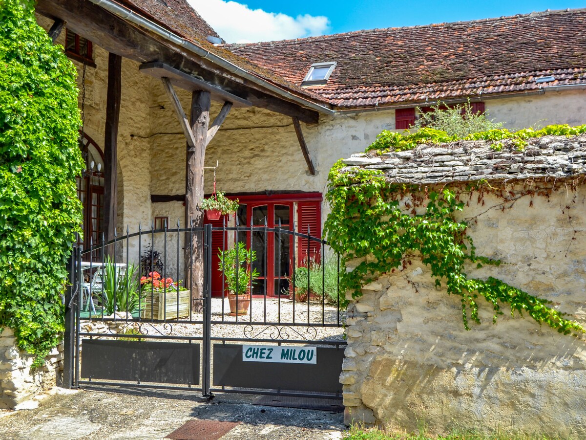The entrance to the rustic cottage features a wrought iron gate labeled 'Chez Milou', surrounded by lush greenery. The stone façade is complemented by red shutters and a flowering garden, creating a welcoming appeal. Sunlight brightens the area, enhancing its charm.