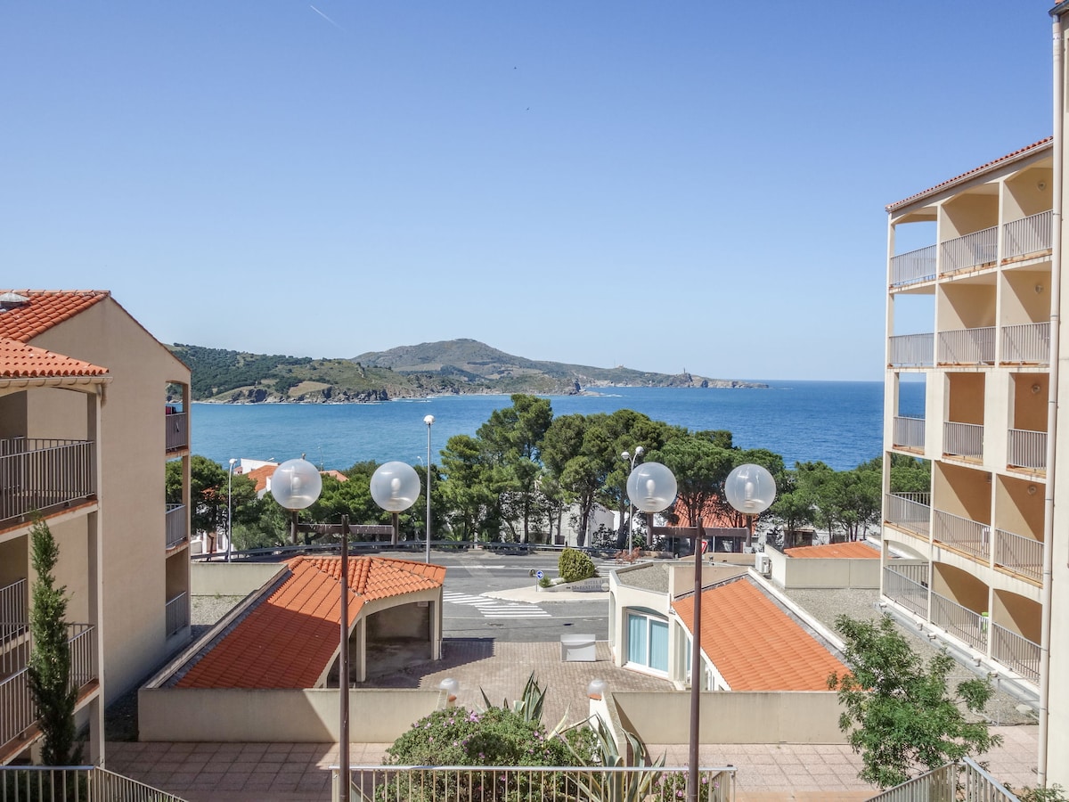 The image showcases a scenic coastal view featuring the sea and distant mountains. The balcony overlooks a well-maintained area with trees and buildings, while bright blue skies contribute to the overall tranquil setting.
