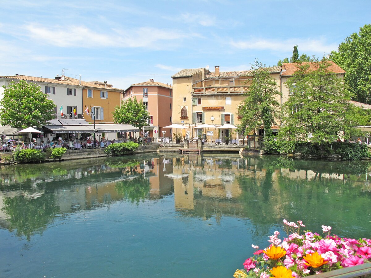 A scenic view of buildings reflecting in a calm body of water. Outdoor dining areas line the shoreline, with tables shaded by umbrellas. Lush greenery surrounds the area, while colorful flowers adorn the foreground, enhancing the tranquil environment.