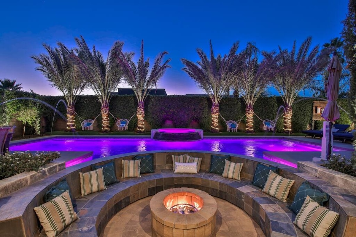 An inviting outdoor space features a circular stone fire pit surrounded by comfortable seating. Lush palm trees are illuminated by soft purple lights, complementing the tranquil saltwater pool in the background. Soft cushions and pillows enhance the relaxing atmosphere.