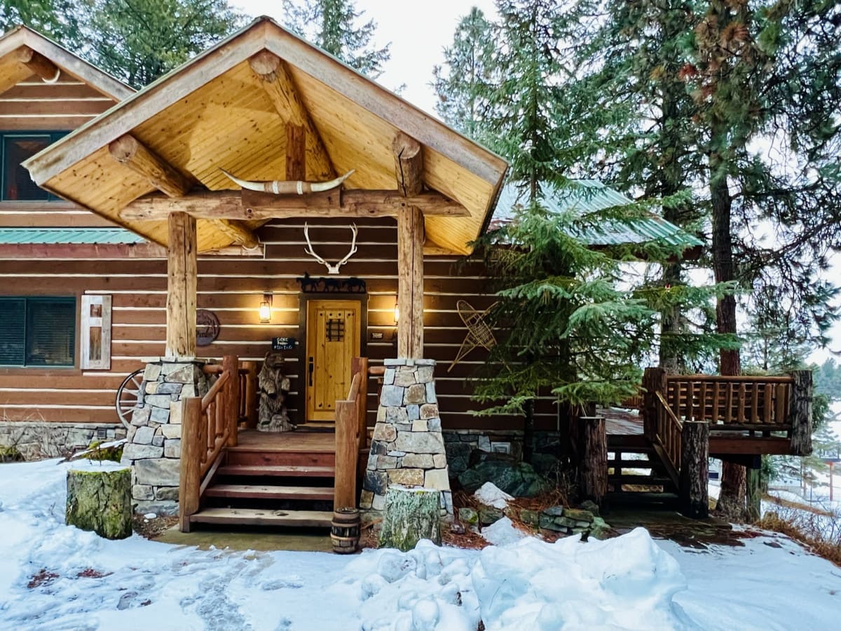 The entrance to the log-style home is framed by tall trees and features a large wooden door complemented by stone accents. A stone staircase leads to the main entry, while a separate staircase provides access to an upper-level balcony. Snow covers the ground, adding to the rustic charm.