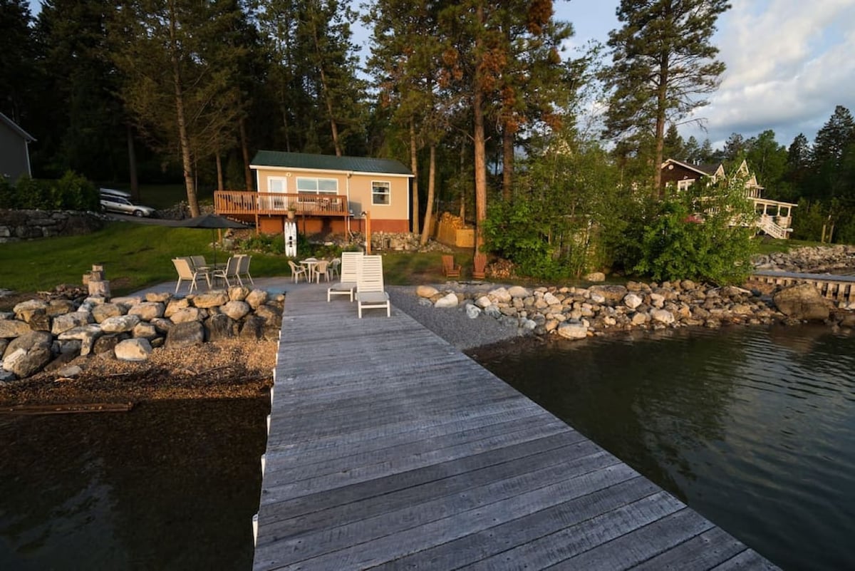 A wooden dock leads to a cottage nestled among trees, showcasing a gradual slope from the lawn to the pebble beach. Multiple lounge chairs are arranged atop the dock, with a backdrop of peaceful water and natural scenery.
