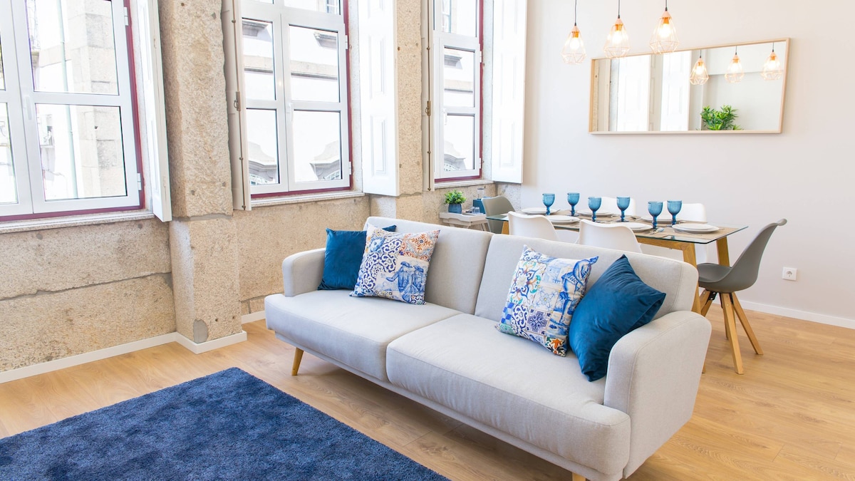 Natural light fills a modern living area, where a light grey sofa adorned with patterned cushions faces a dining table set for six. Large windows with white frames reveal views of the street below, while a stylish mirror and pendant lights enhance the spacious feel.