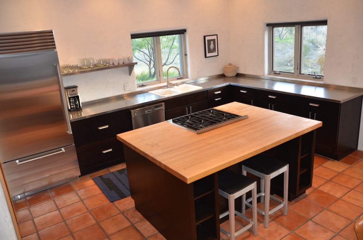 A modern kitchen is equipped with premium appliances, including a stainless steel refrigerator and a gas range. The spacious island features seating for three. Natural light filters through two windows, highlighting the tile floor and sleek counters, creating an inviting atmosphere for cooking.